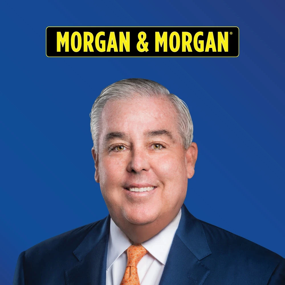 Portrait of a smiling man in a blue suit and orange tie in front of a solid blue background with the Morgan & Morgan logo above him. Professional image representing the face of the law firm.