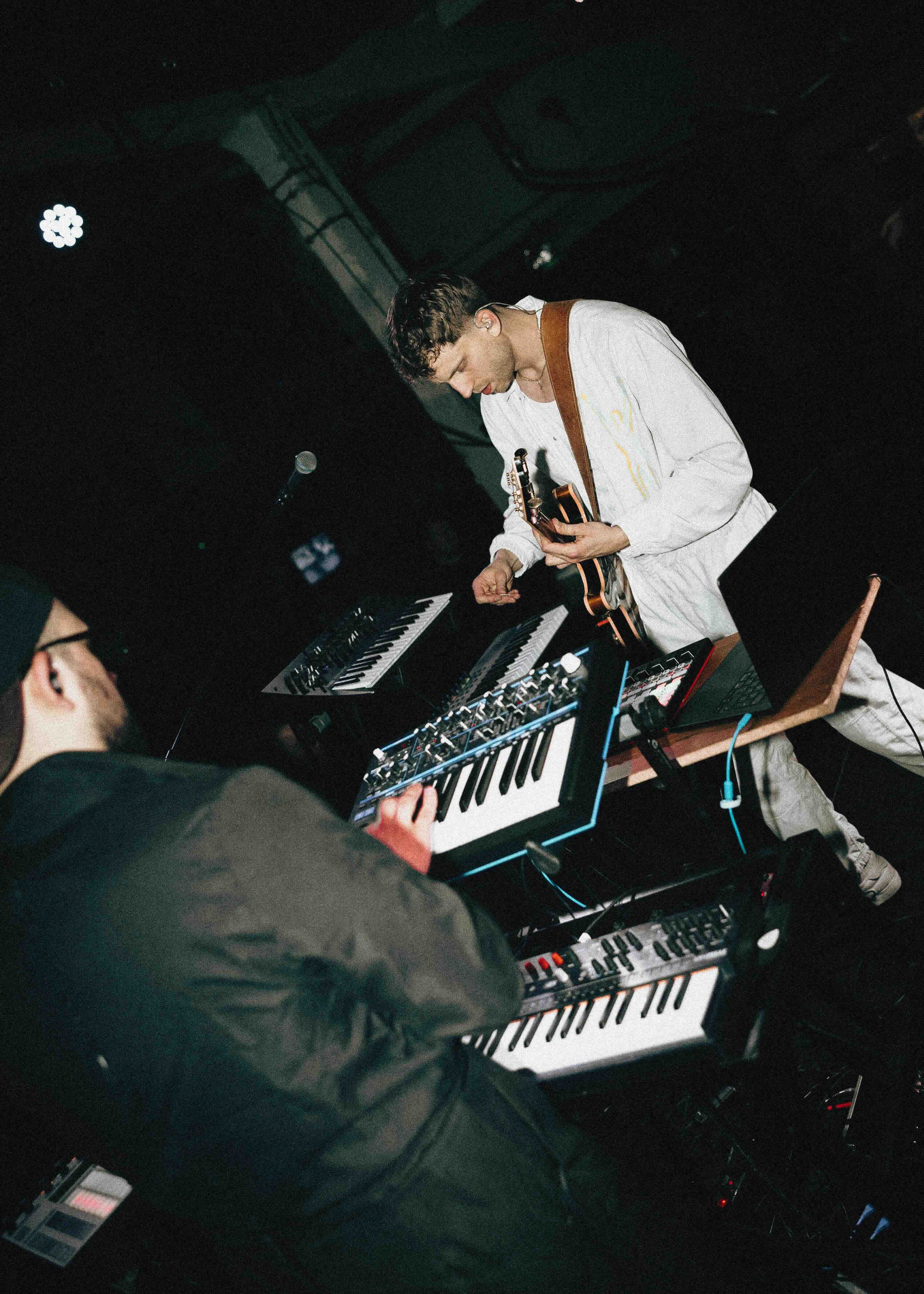 A musician in a white outfit playing guitar while another in black stands with their back to the camera playing a synthesizer, shot on a dark stage