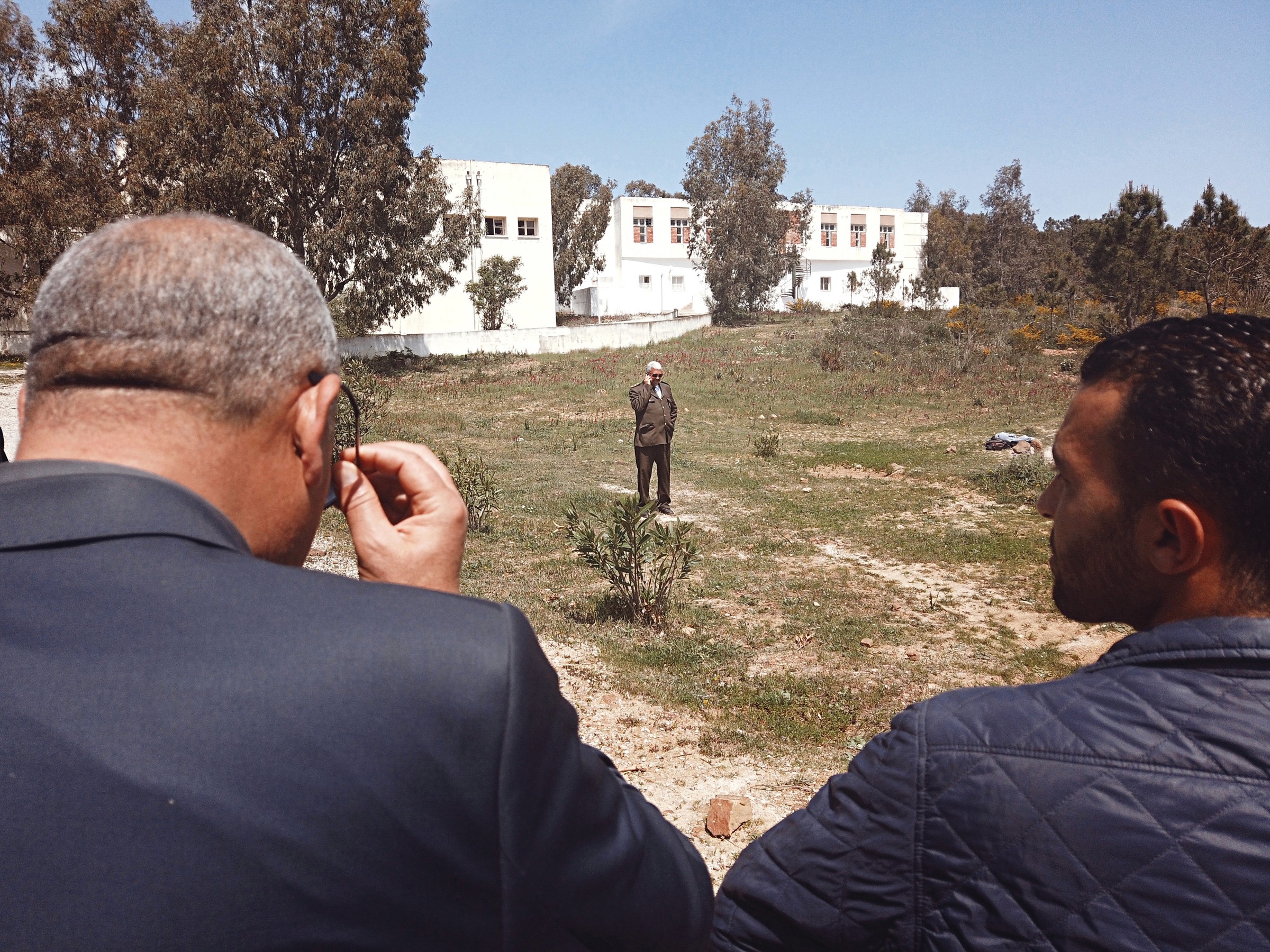 Photo au dessus des épaules de deux hommes montrant un homme au loin au téléphone dans un terrain vague à l'extérieur d'une école en Tunisie