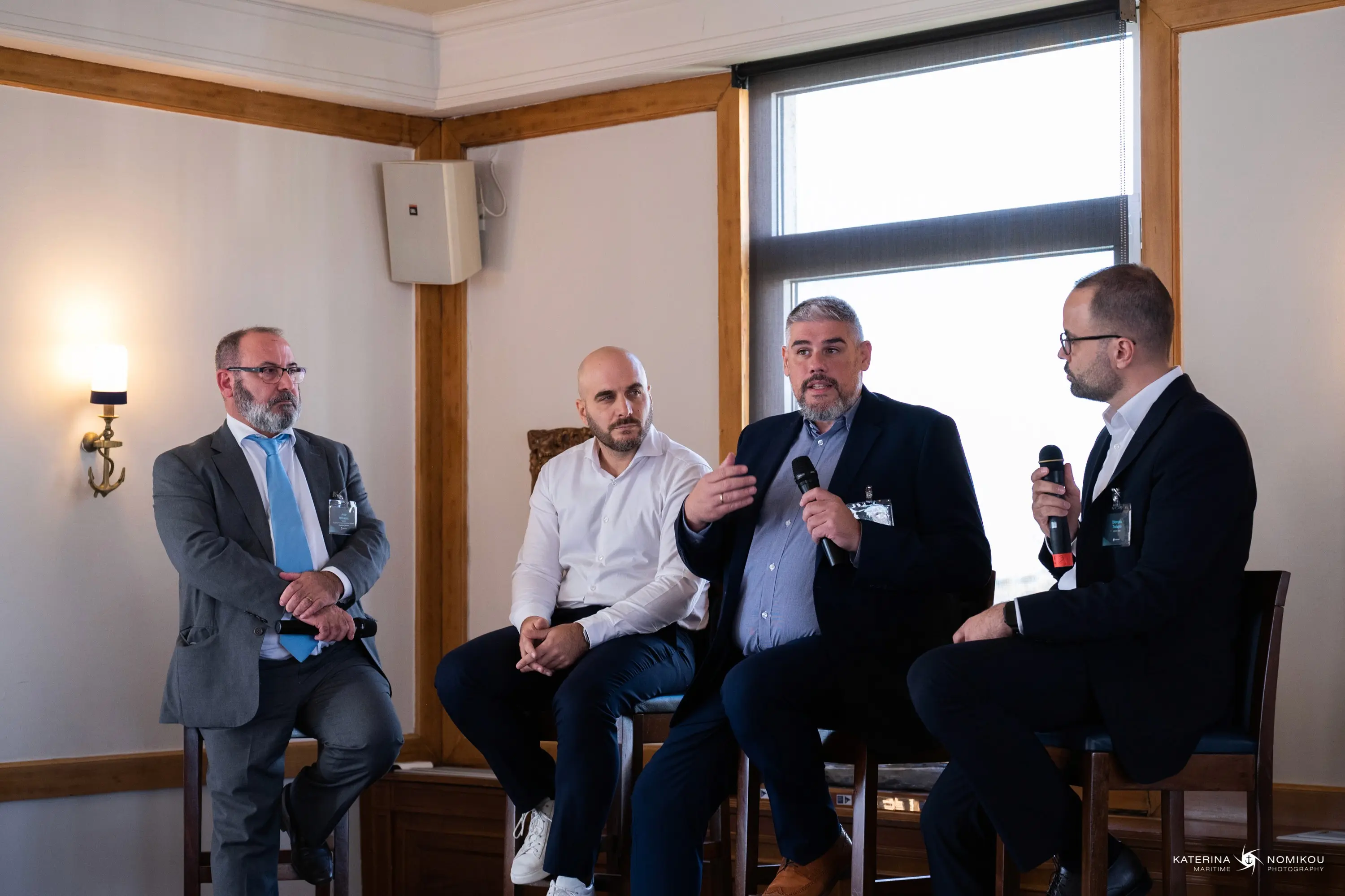 A panel of four men engaged in discussion, seated on stools in a bright, airy room.