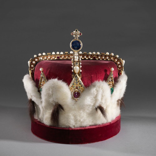 An ornate red velvet crown with pearls, gemstones, and a fur trim, set against a dark background.