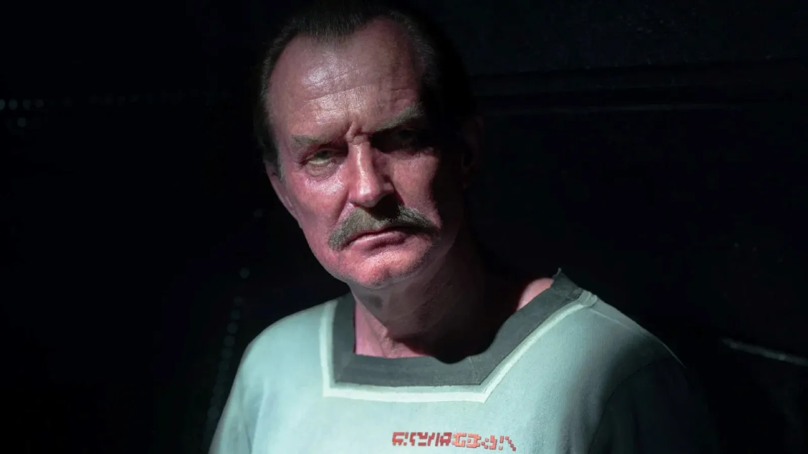 The image features a man in low lighting wearing a light green shirt with orange text, displaying a serious expression, against a dark background, creating a mysterious and intense atmosphere.