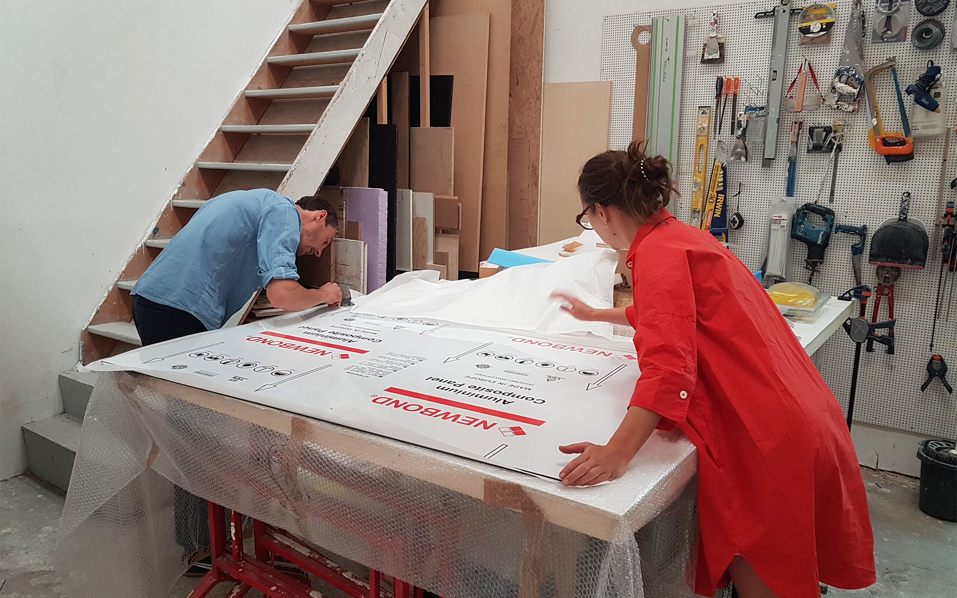 Deux personnes travaillent ensemble sur une grande table dans un atelier, positionnant une large plaque de panneau composite en aluminium de la marque « NEWBOND ». Un homme en chemise bleue est à gauche, concentré sur le bord du panneau, tandis qu'une femme en chemise rouge vif se tient à droite pour l'aider à l'aligner. La table est recouverte de papier bulle et, derrière eux, un mur est organisé avec divers outils à main suspendus sur un panneau perforé.