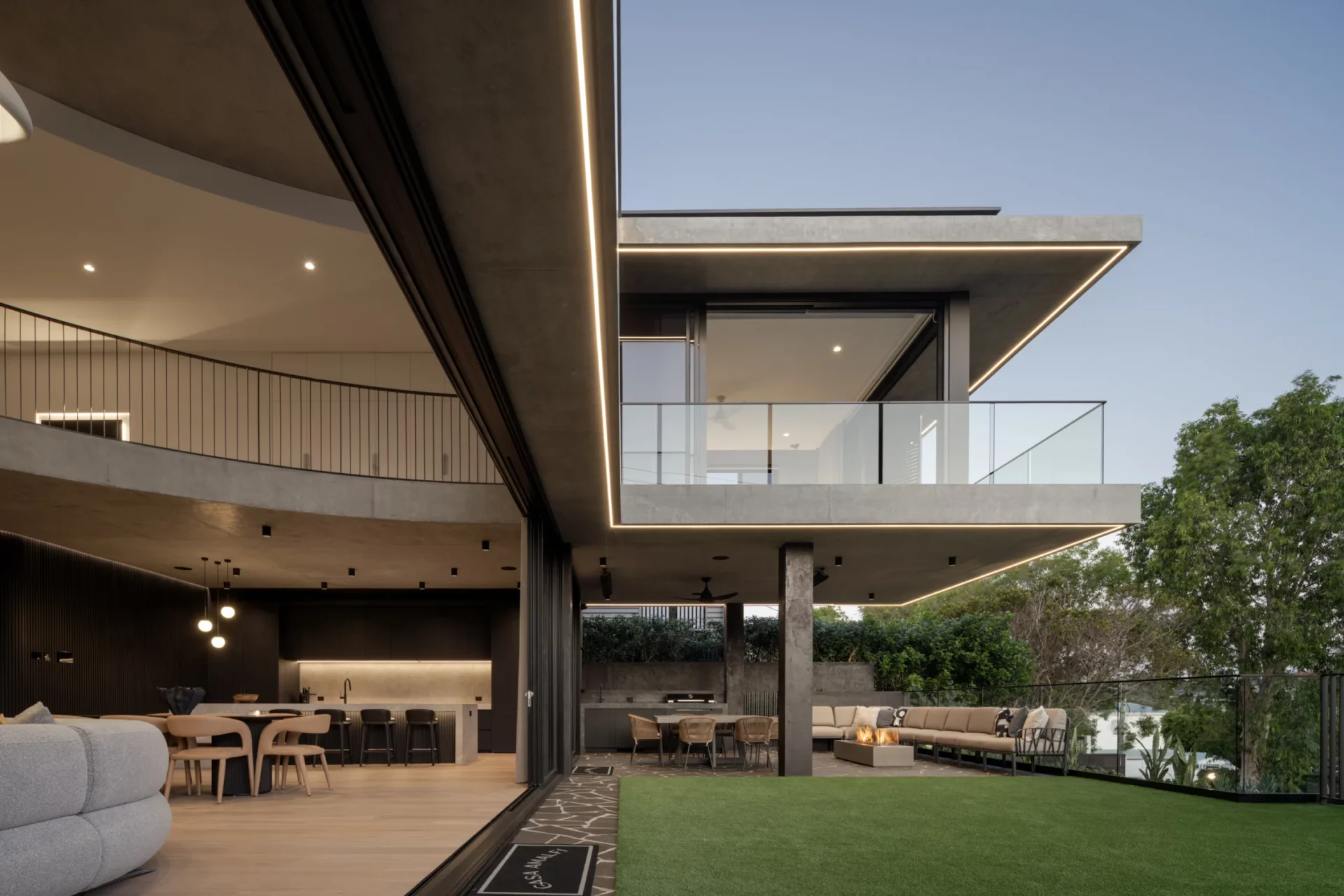 An open office space with large windows and industrial decor. A person sits on a bench working on a laptop, surrounded by lush green plants and sleek furniture.