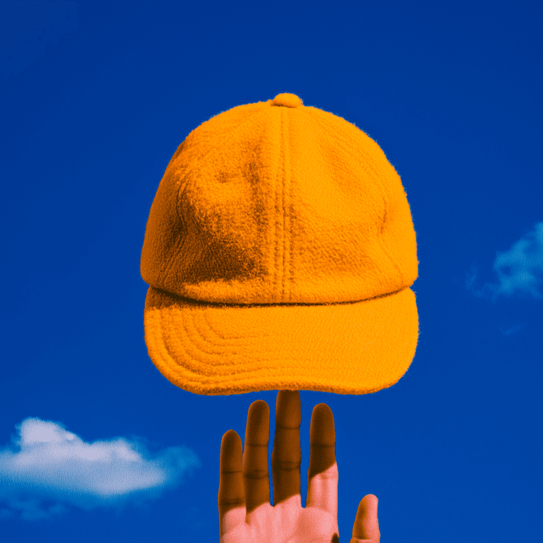Bright orange textured cap balanced on fingertips against a vivid blue sky with scattered clouds.