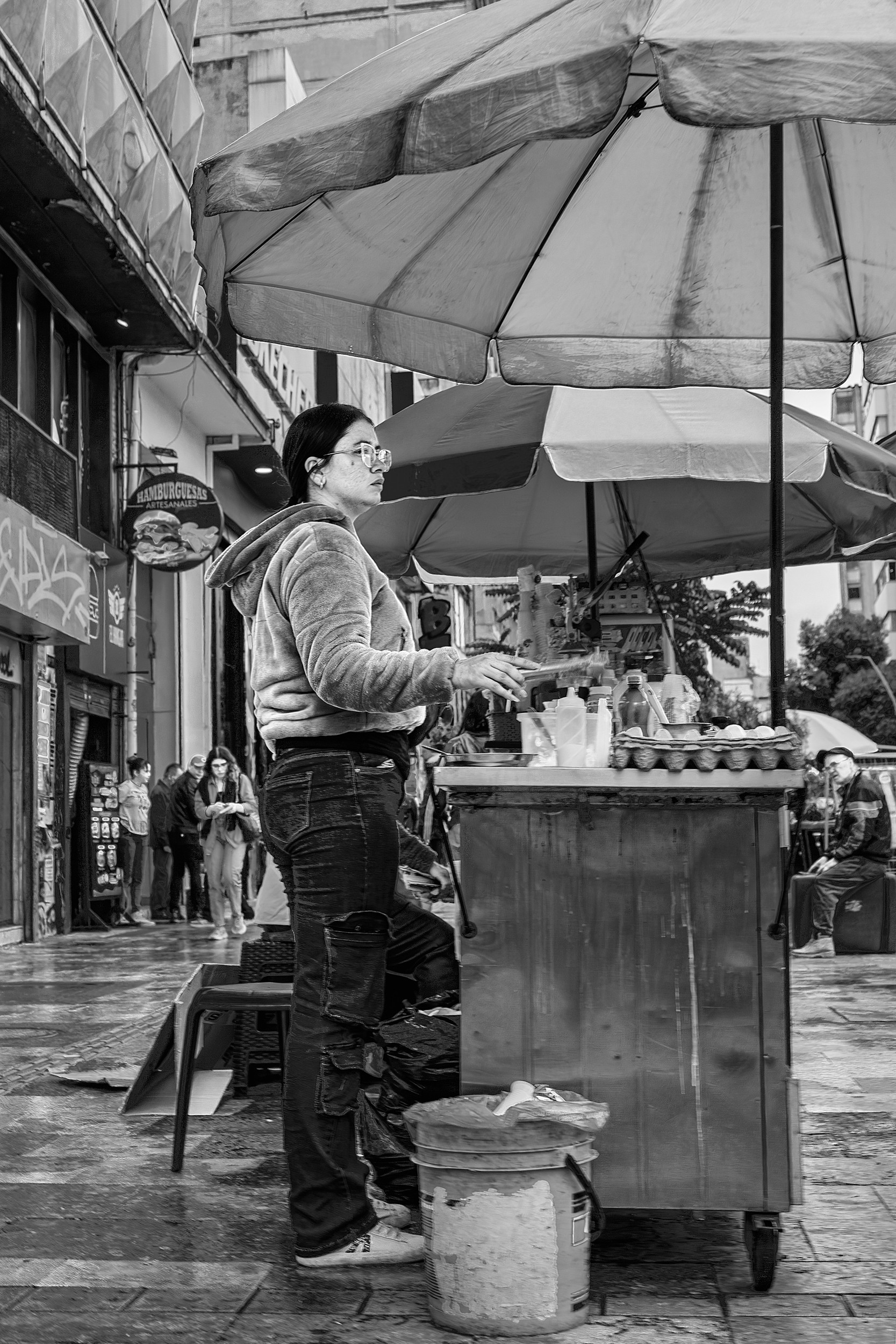 A woman stands behind a small food cart topped with condiments and eggs, working under large umbrellas on a busy sidewalk in Bogotá.