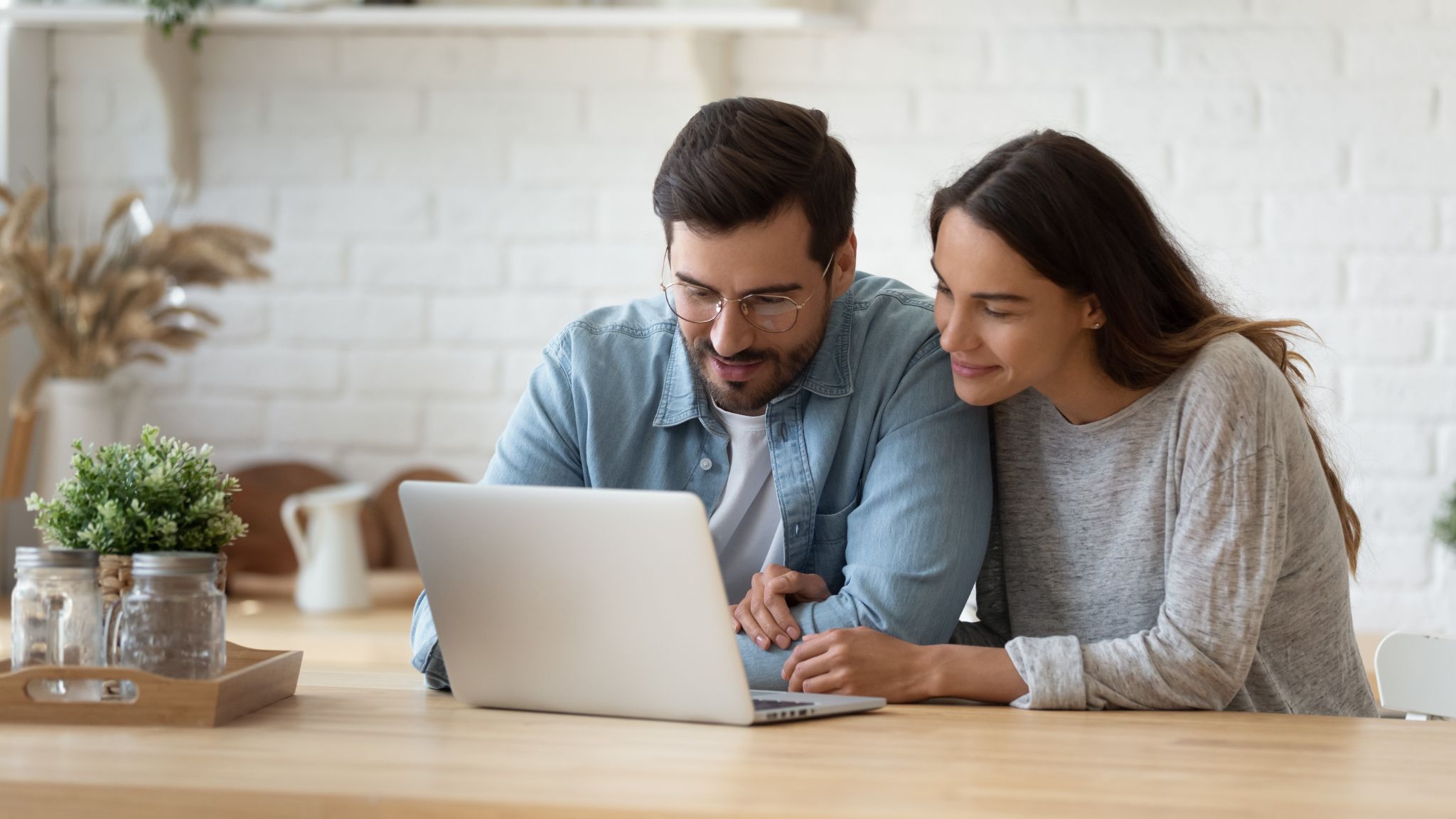 Ein Mann mit Brille und eine Frau schauen gemeinsam auf einen Laptop auf einem Holztisch.