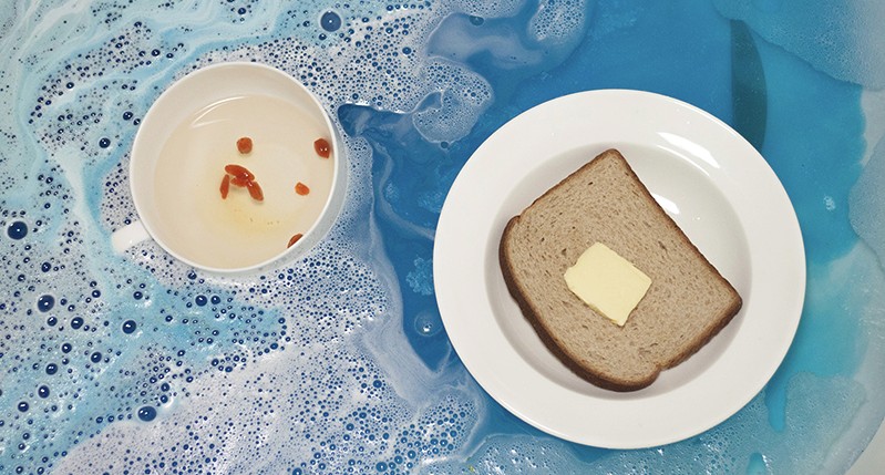 A photo of a plate with a piece of bread and butter, a cup with some water and some red seeds. Both of them are placed in a blue colored water.