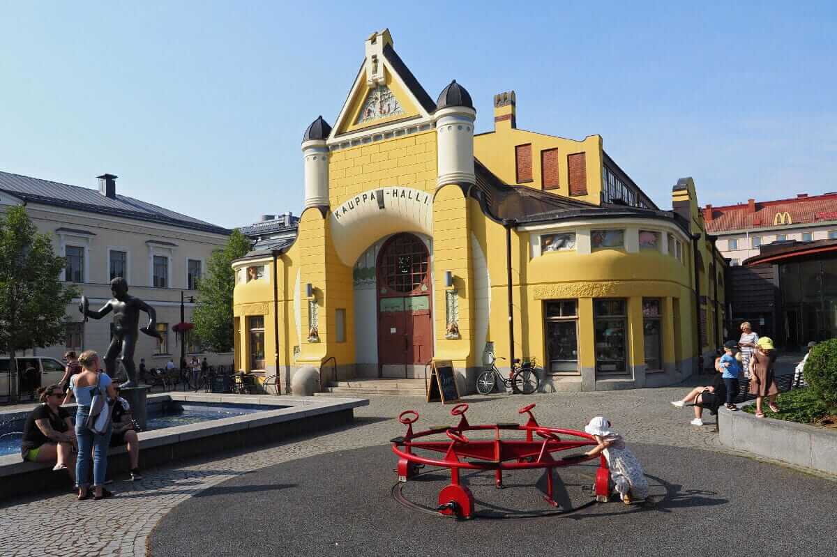 A man wearing glasses and a blazer working at a desk, writing in a notebook beside a computer monitor in a modern office.