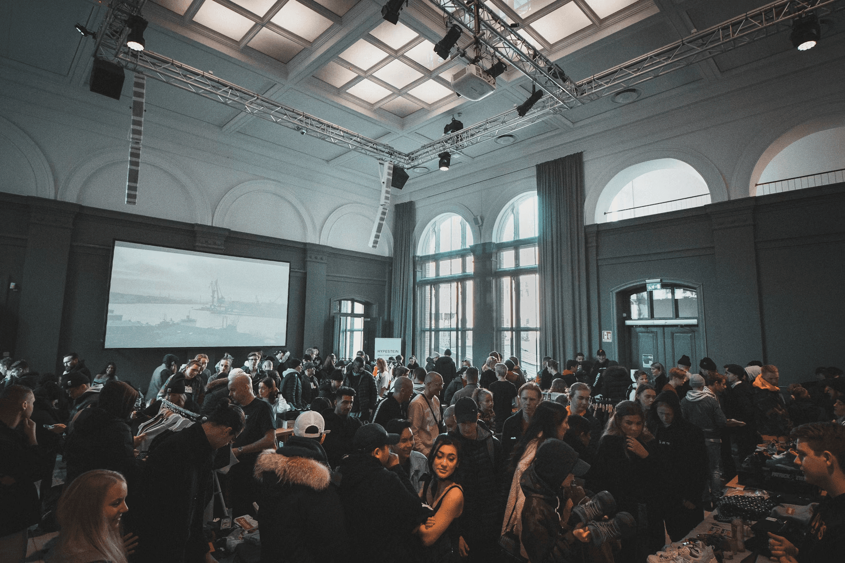 A crowded conference hall with stands and a presentation in motion. Photo by Jakob Dalbjörn on Unsplash
