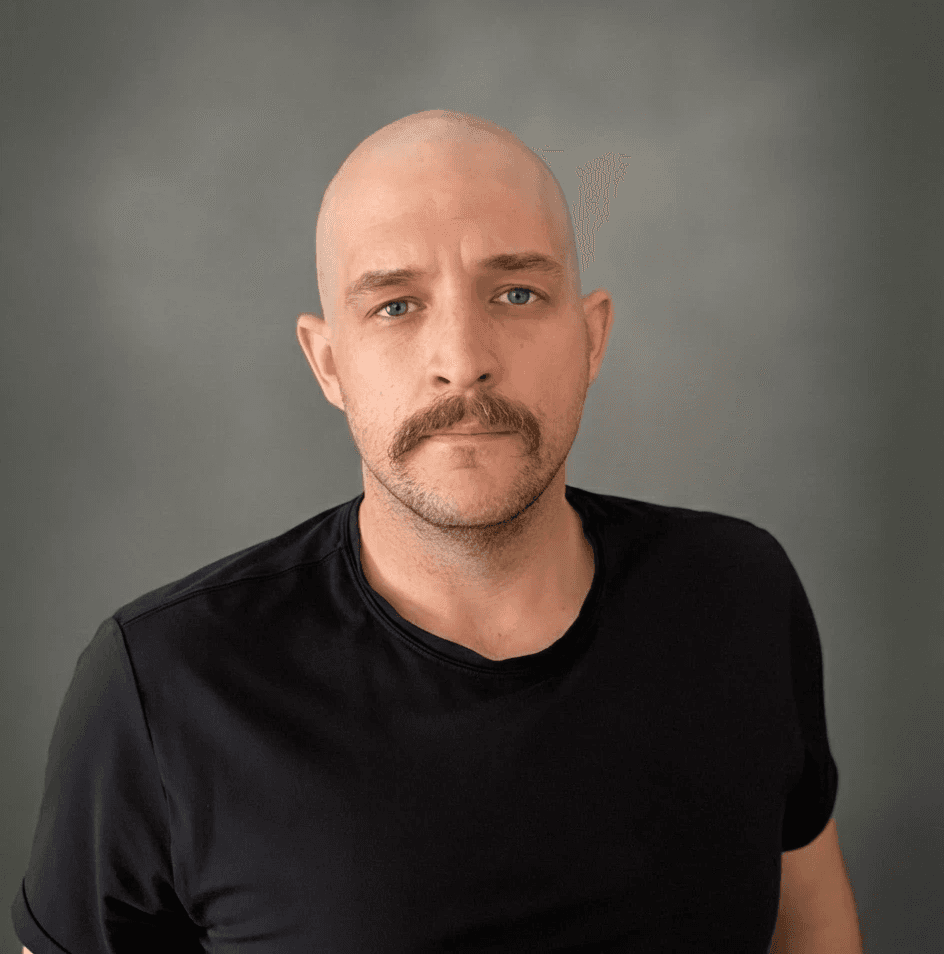 Um homem com bigode e cabeça raspada, usando uma camiseta preta, está em pé diante de um fundo cinza texturizado.