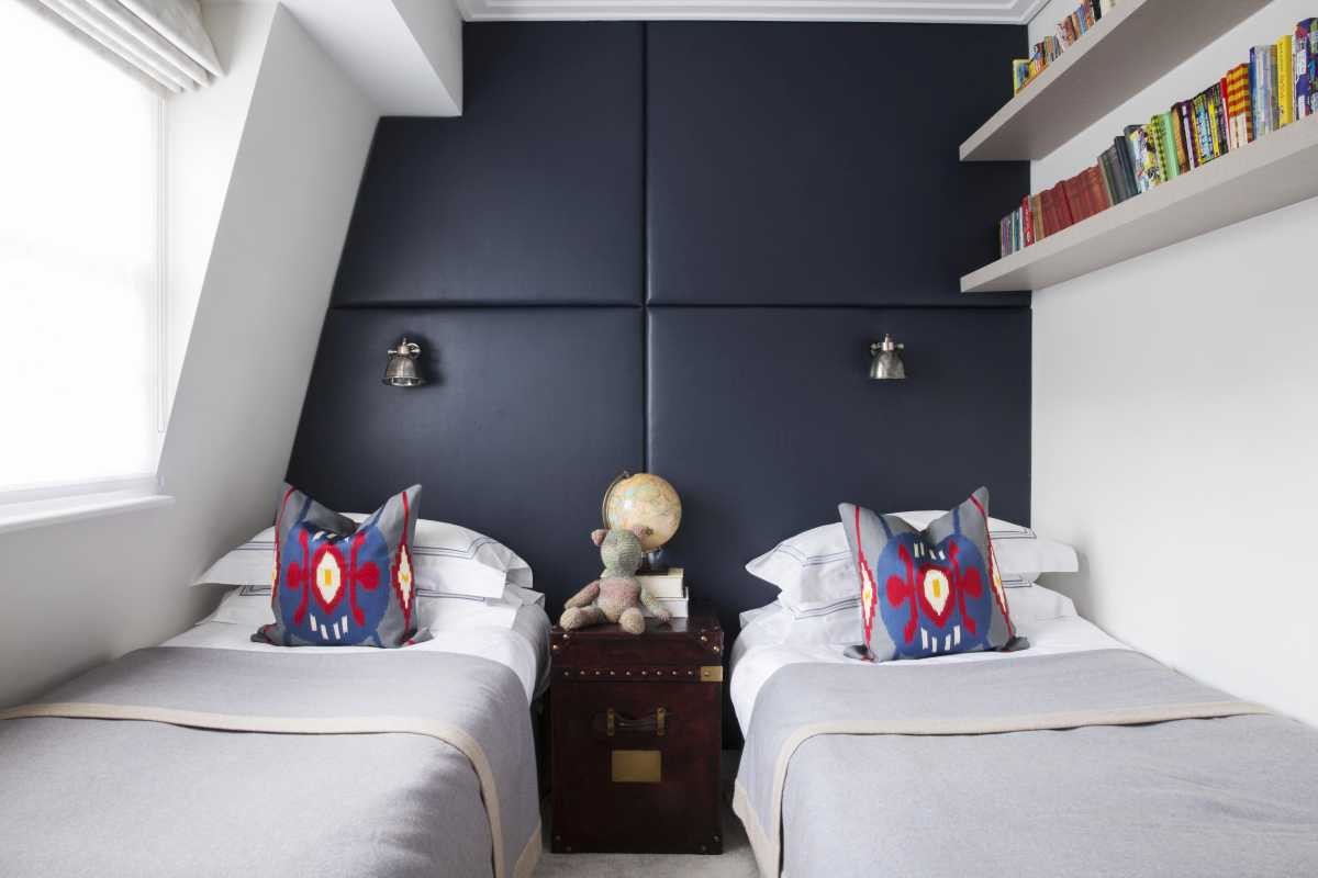 Bespoke twin bedroom layout created by Lymington interior designers, featuring a dark navy upholstered wall panel, wall-mounted reading lights, and floating bookshelves.