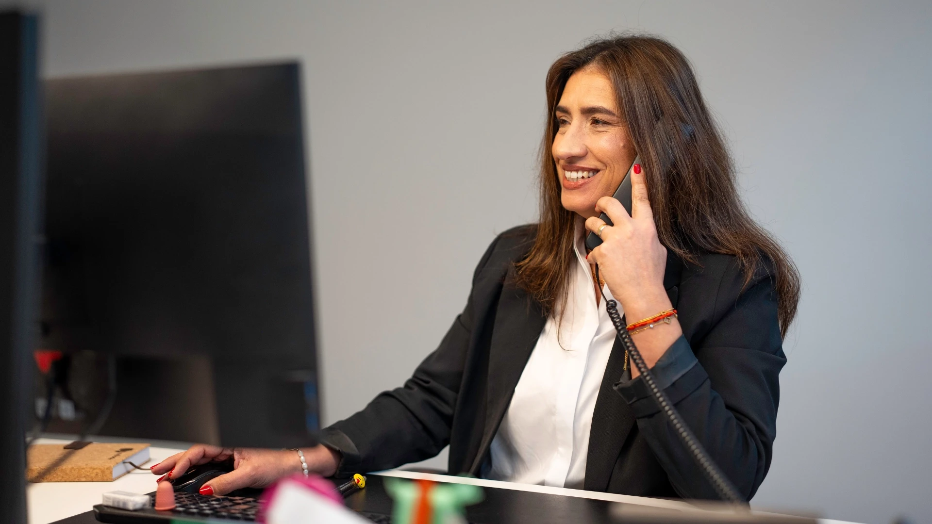 Mulher de negócios sorridente usando telefone e computador em ambiente de escritório, transmitindo profissionalismo e satisfação no trabalho.