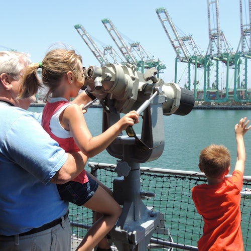 A child looks through binoculars on a dock, assisted by an adult. Another child waves towards visible cranes in the background.