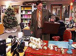Dwight Schrute from The Office, wearing a coat, holding a briefcase. A Christmas tree and wrapped presents are in the office.
