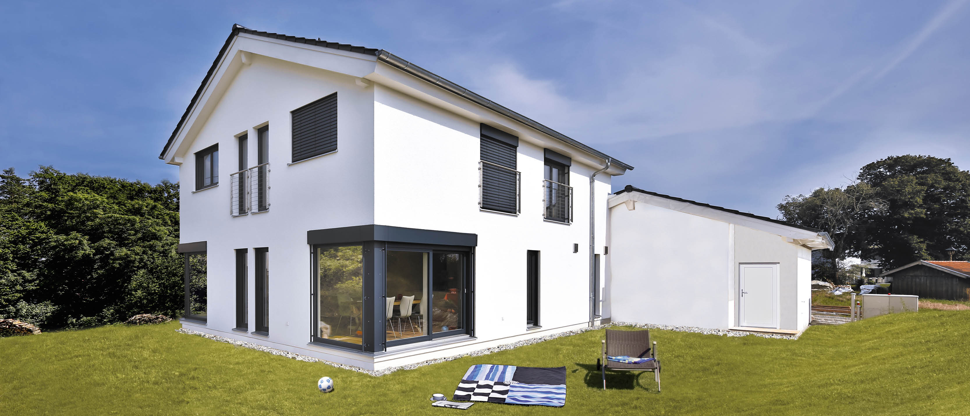 Moderne weiße Villa mit großen Fenstern, Balkonen und Rollos, grasbewachsener Garten mit Liegestuhl, Decke und Ball.