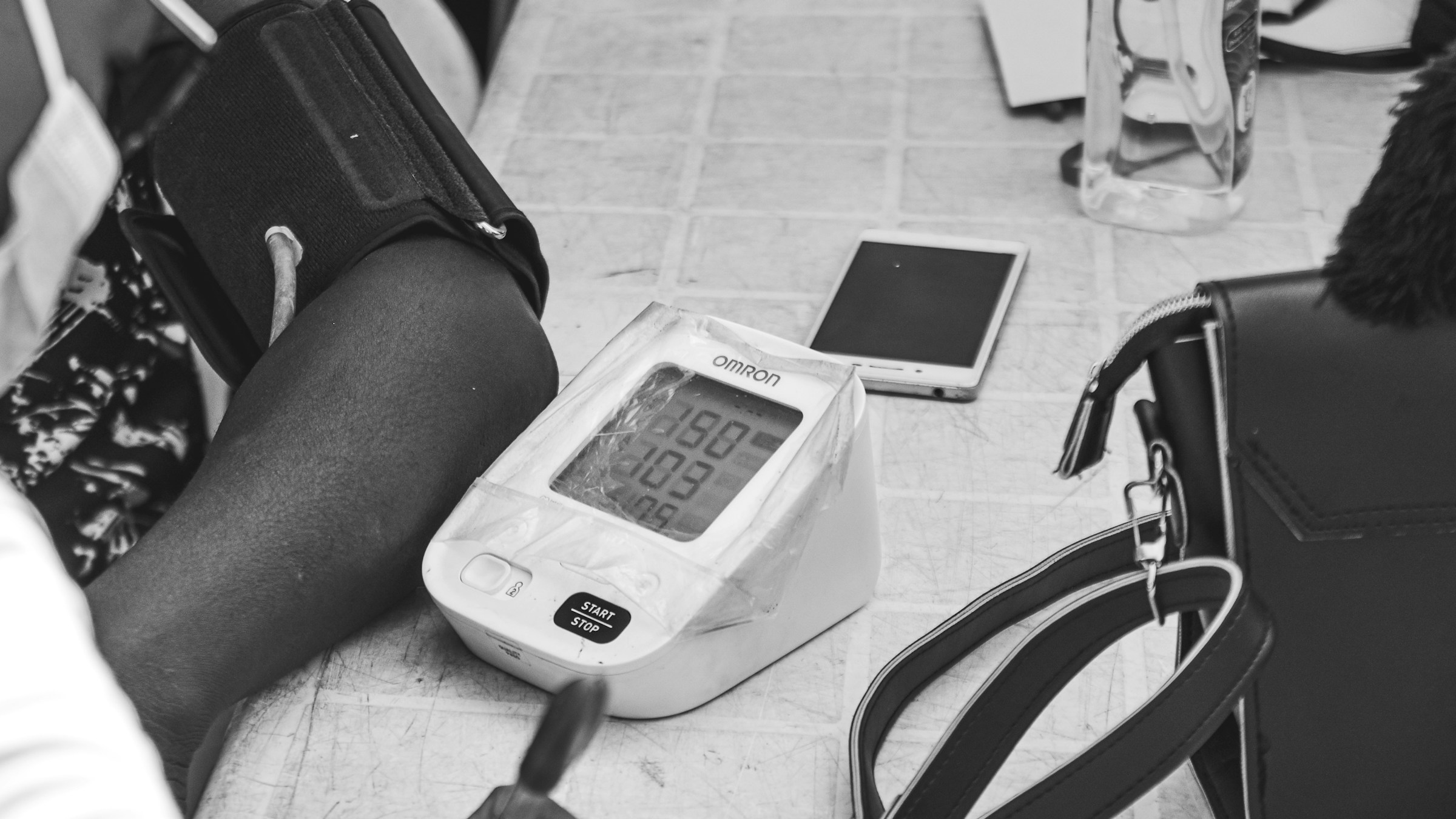 A person's arm resting on a tiled surface with a digital home blood pressure monitor cuff wrapped around the upper arm mid-measurement — a black-and-white documentary photograph of the routine at-home screening moment central to catching diabetes complications early