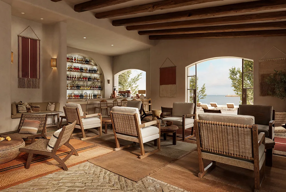 Einladende Hotel-Lounge im mediterranen Stil mit freiliegenden Deckenbalken, gemütlichen Sesseln aus Holz und Geflecht sowie einer gut sortierten Bar im Hintergrund. Große Rundbogenfenster öffnen den Raum zum Meer.