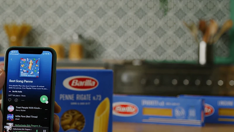 Hand holding smartphone displaying Barilla 'Best Song Penne' Spotify playlist in kitchen with Barilla Penne Rigate No.73 pasta boxes in background, Playlist Timer campaign lifestyle shot