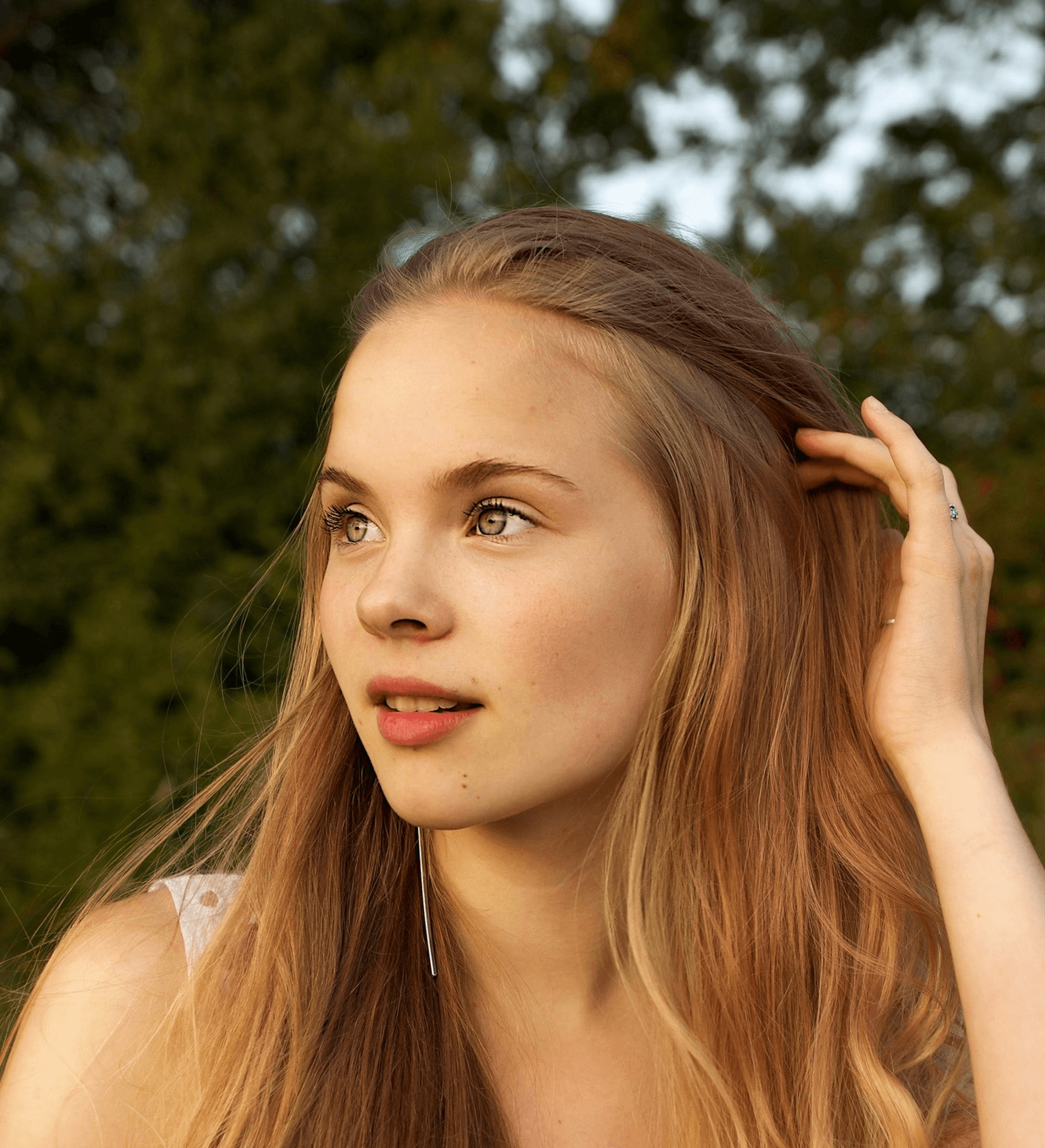 Person with long light brown hair outdoors touching hair, with green foliage background