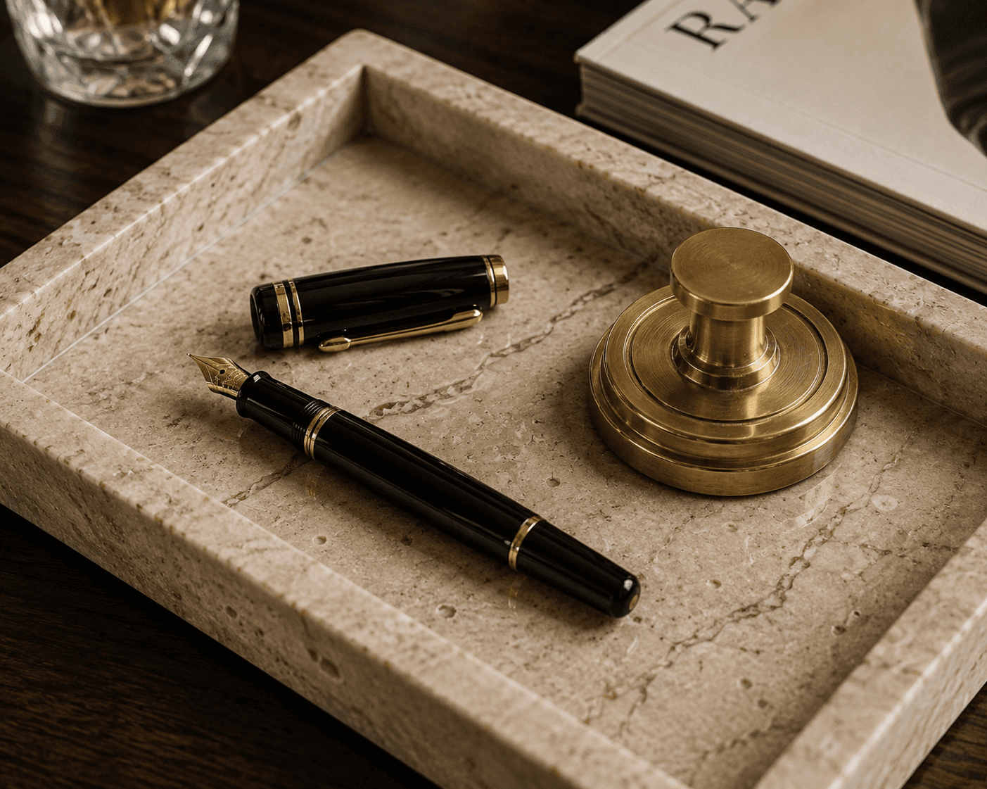 Close-up shot of high-end desk accessories resting in a beige marble valet tray. The tray contains a classic black Montblanc fountain pen with its cap unscrewed to reveal a gold nib, alongside a heavy, circular brass paperweight. A corner of an art book and a crystal glass are partially visible on the dark wood surface nearby.