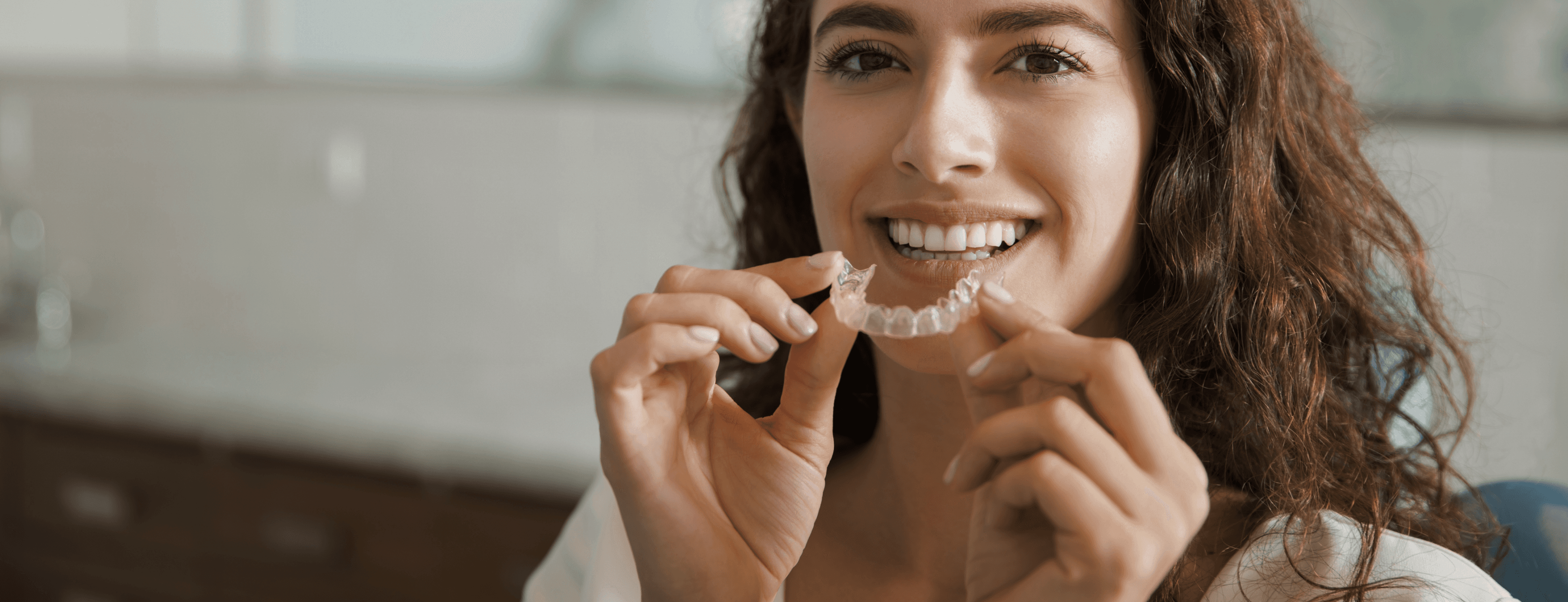 A smiling woman holding a clear aligner, highlighting the benefits of using aligners for straightening teeth.