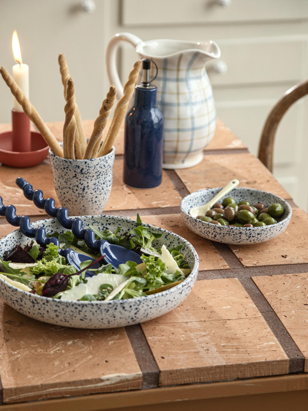 Grand saladier en céramique mouchetée bleu et blanc garni de salade verte avec couverts bleus, accompagné d'olives et de grissinis sur table carrelée