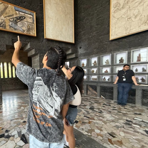 Dalawang tao ang nagtuturo sa naka-frame na sining sa dingding ng gallery, habang may isang tao na nakatayo malapit sa isang display ng maliliit na eskultura.