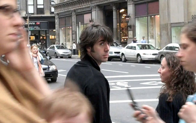 A man with medium-length brown hair, wearing a black shirt, stands in the center of a city street. He is looking to his right. Behind him, a woman with long, dark brown hair and a black top is visible. To his left, a person with glasses and a tan garment is partially visible. Other people can be seen in the background, and cars are parked along the street. The scene is set near a building with large windows.