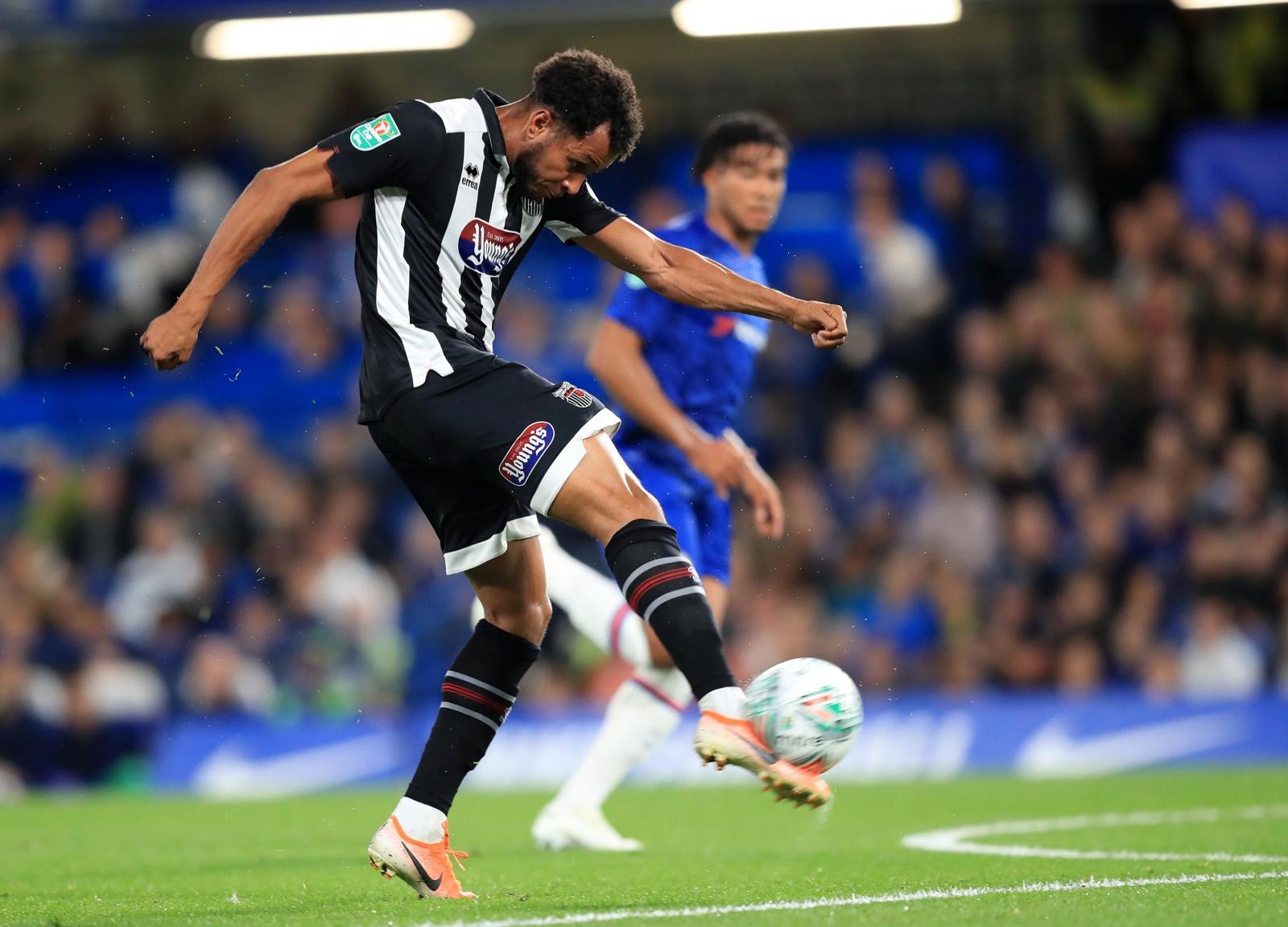 A football player in a black and white striped jersey powerfully kicks a ball during a match, with a blurred opponent in blue and a cheering crowd in the background.