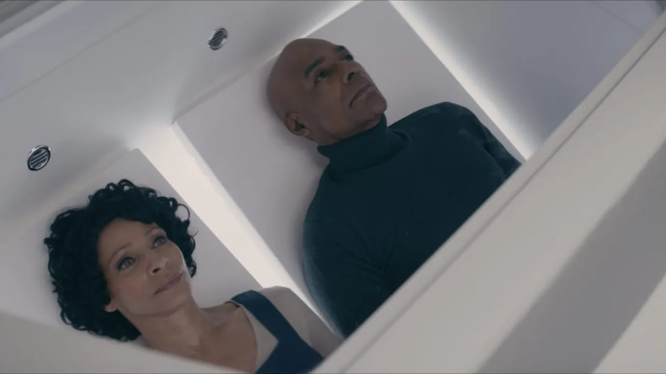 Two individuals lie side by side in a futuristic, illuminated white chamber, suggesting a sci-fi setting or advanced technology environment.