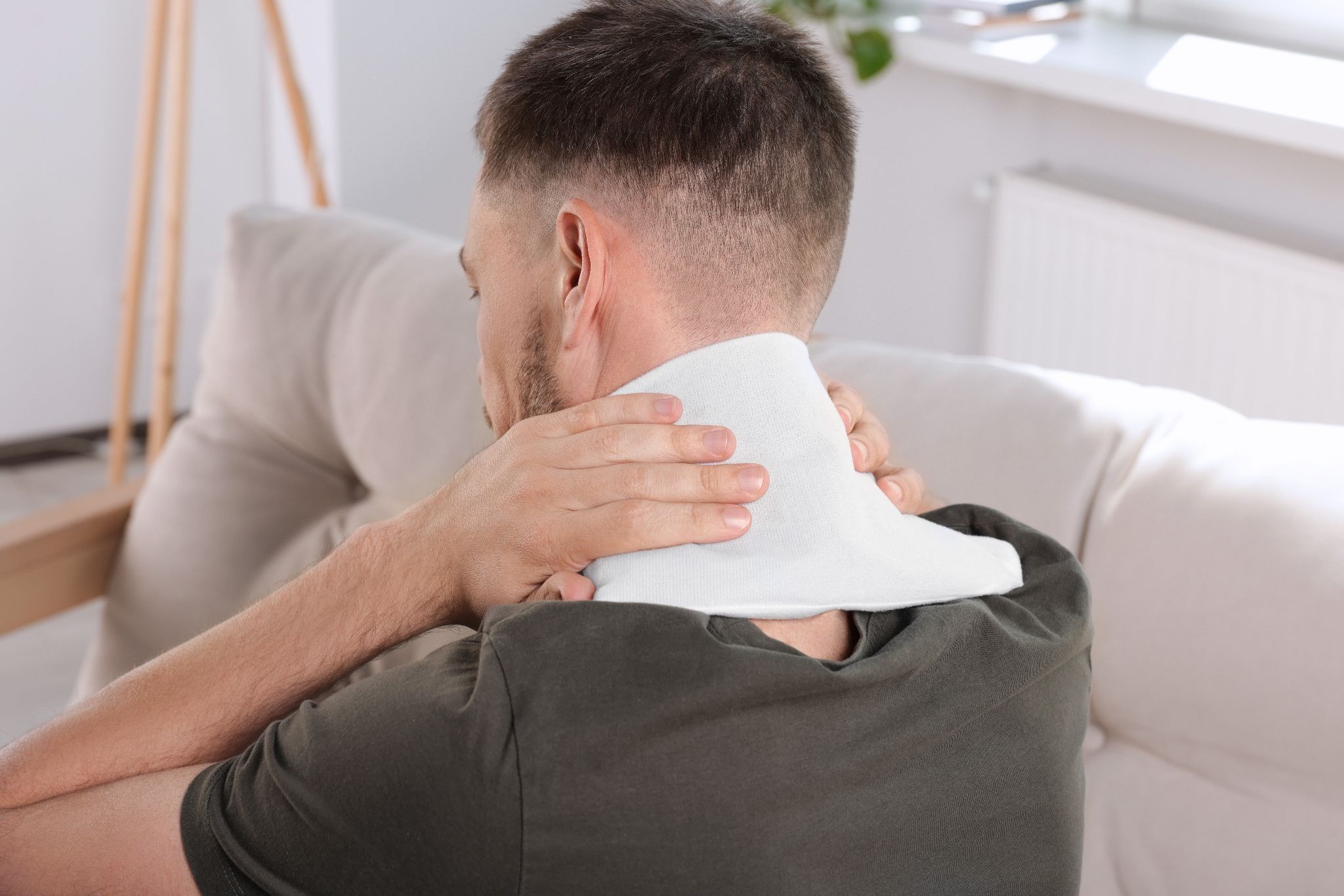 A man seen from behind sits on a couch while holding a white heating pad or compress to his neck.