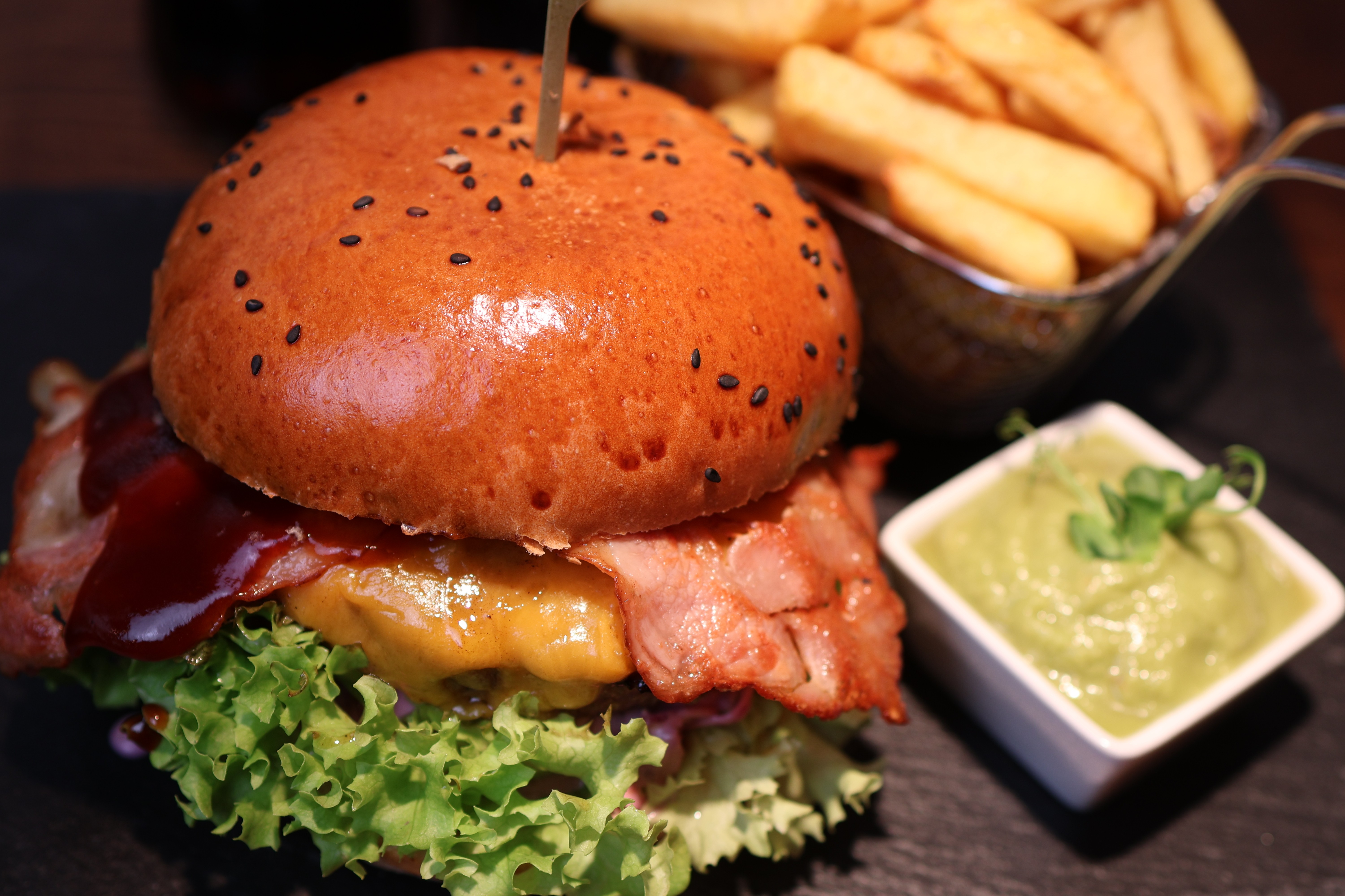Bacon-Cheeseburger im kAlps mit Sesambrötchen, geschmolzenem Käse, Salat, knusprigem Speck, Pommes und grünem Dip.