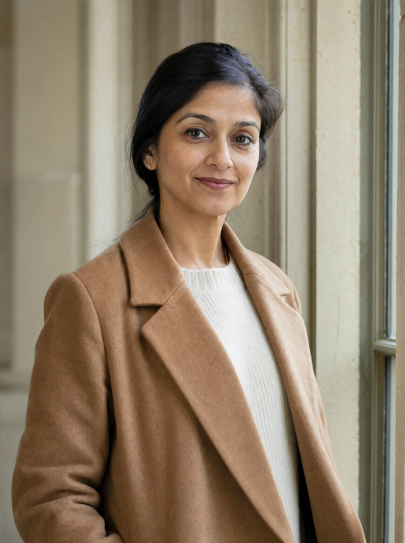 Priya Nair, partner, in a camel coat beside a tall window