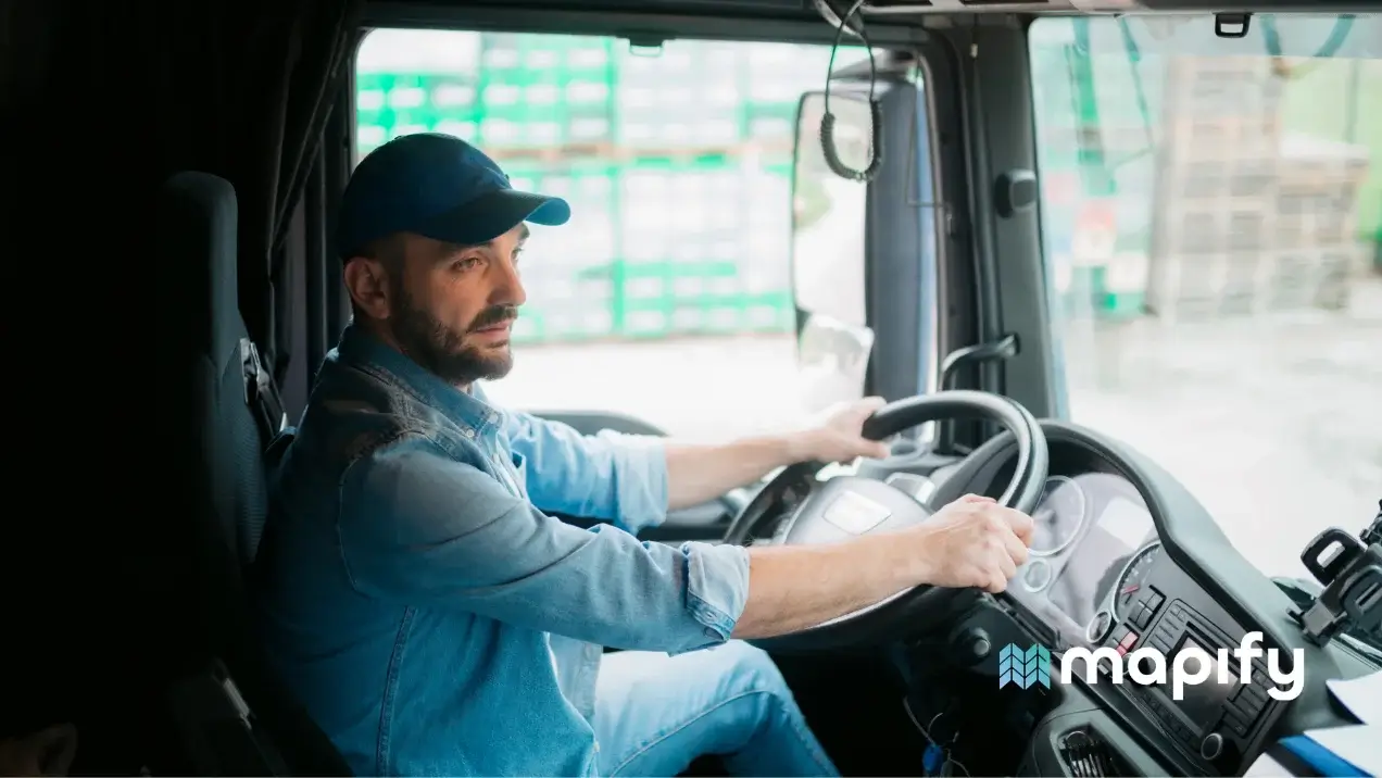 A man in a denim shirt and cap is driving a truck, focused on the road. The interior is dimly lit, creating a calm and professional atmosphere.