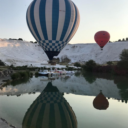 Isang asul-at-puting mainit na air balloon na may repleksyon sa isang lawa, habang may isa pang pulang balloon sa background sa puting lupain.
