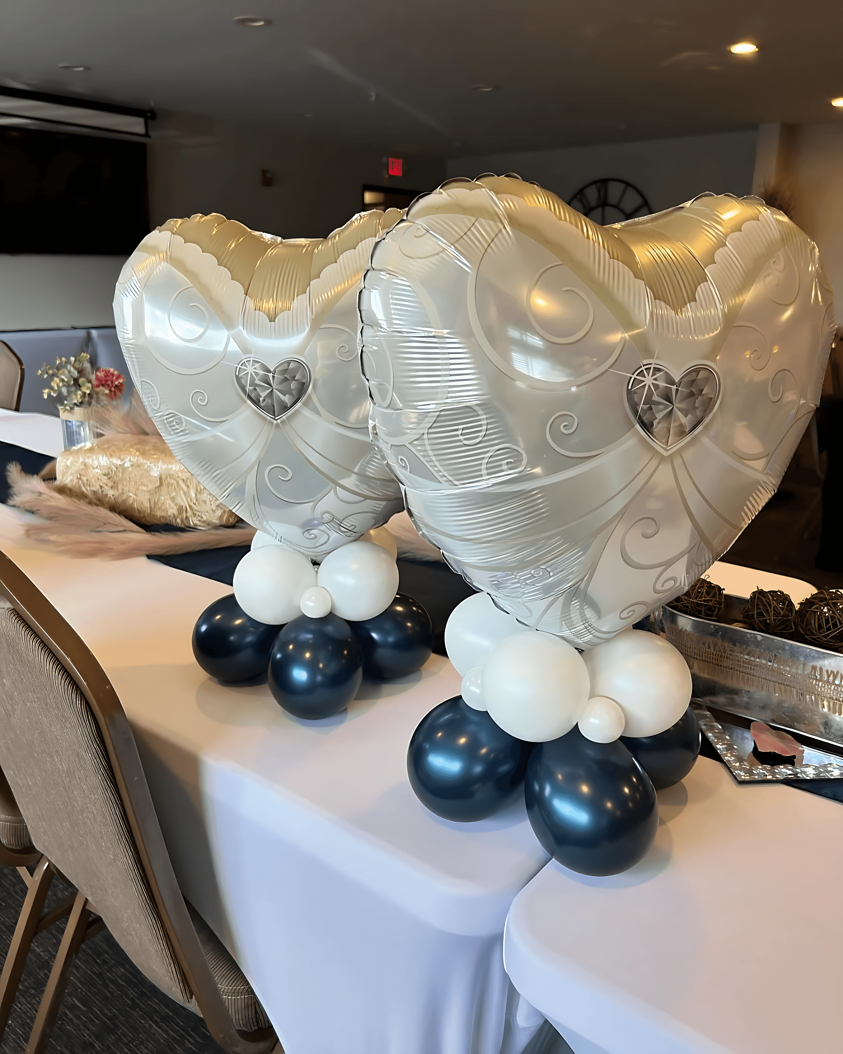 Pair of gold and white heart-shaped balloon centerpieces with blue bases displayed on round tables for a celebration by Be Bold Balloons
