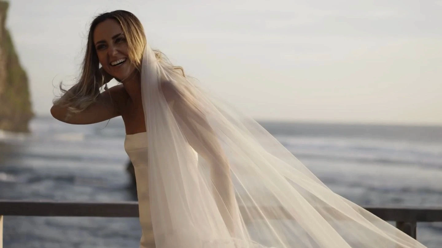 A smiling bride in a white wedding dress with a long veil stands on a balcony overlooking the ocean, with rocky cliffs in the background, under a clear sky.