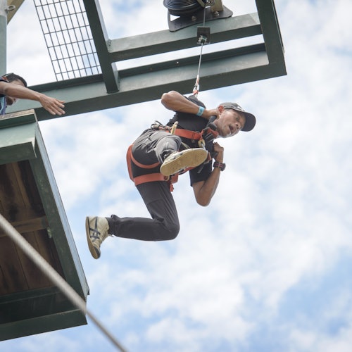 Person in a harness and helmet ziplining from a platform, with a clear blue sky in the background.