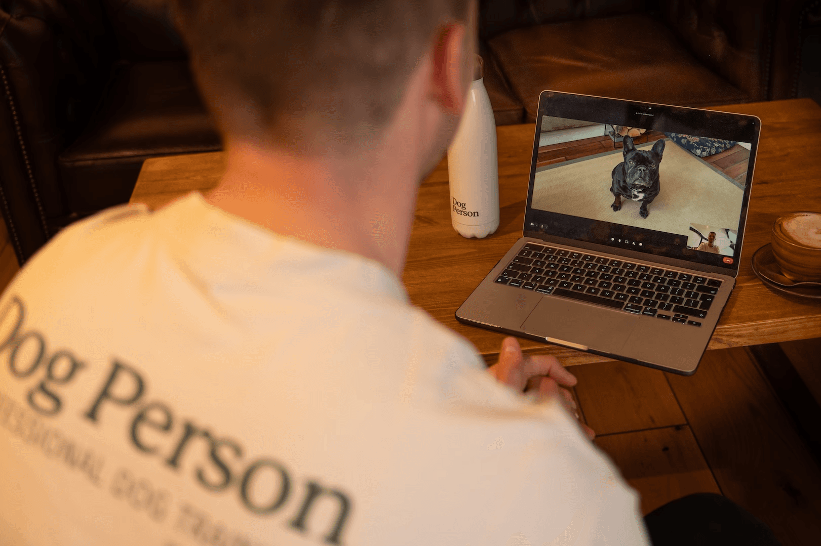 Scott (Dog Person) is sat at the laptop on a video call (wearing branded t-shirt). The laptop screen shows a French Bulldog sat down on a carpet. Beside the laptop is a coffee and a Dog Person branded water bottle
