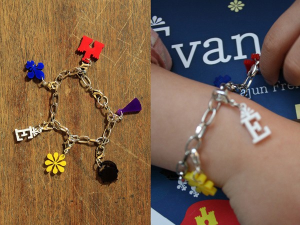 The image shows a silver chain bracelet with colorful letter charms. The left side displays a close-up of the bracelet on a wooden surface, showcasing the chain and charms: a red letter “H,” a blue flower, a yellow flower, a purple flower, and a black flower. The right side shows a hand wearing a similar bracelet with a white letter “E” charm and a yellow flower charm. The background includes a blue surface with partial white text visible.