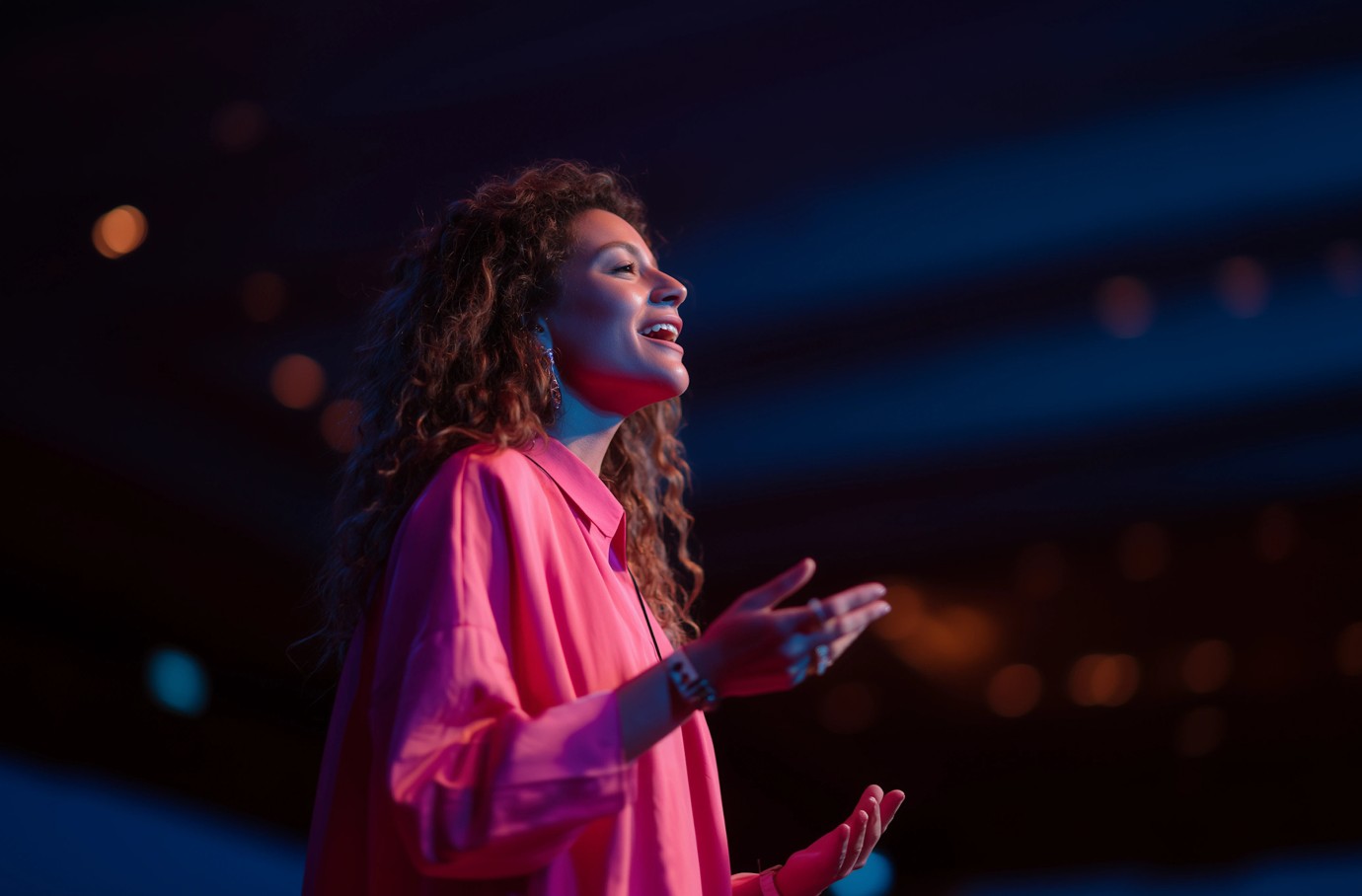 A person in a bright pink shirt stands under dramatic stage lighting, passionately speaking with expressive gestures, capturing an atmosphere of dynamic public speaking or a live performance event.