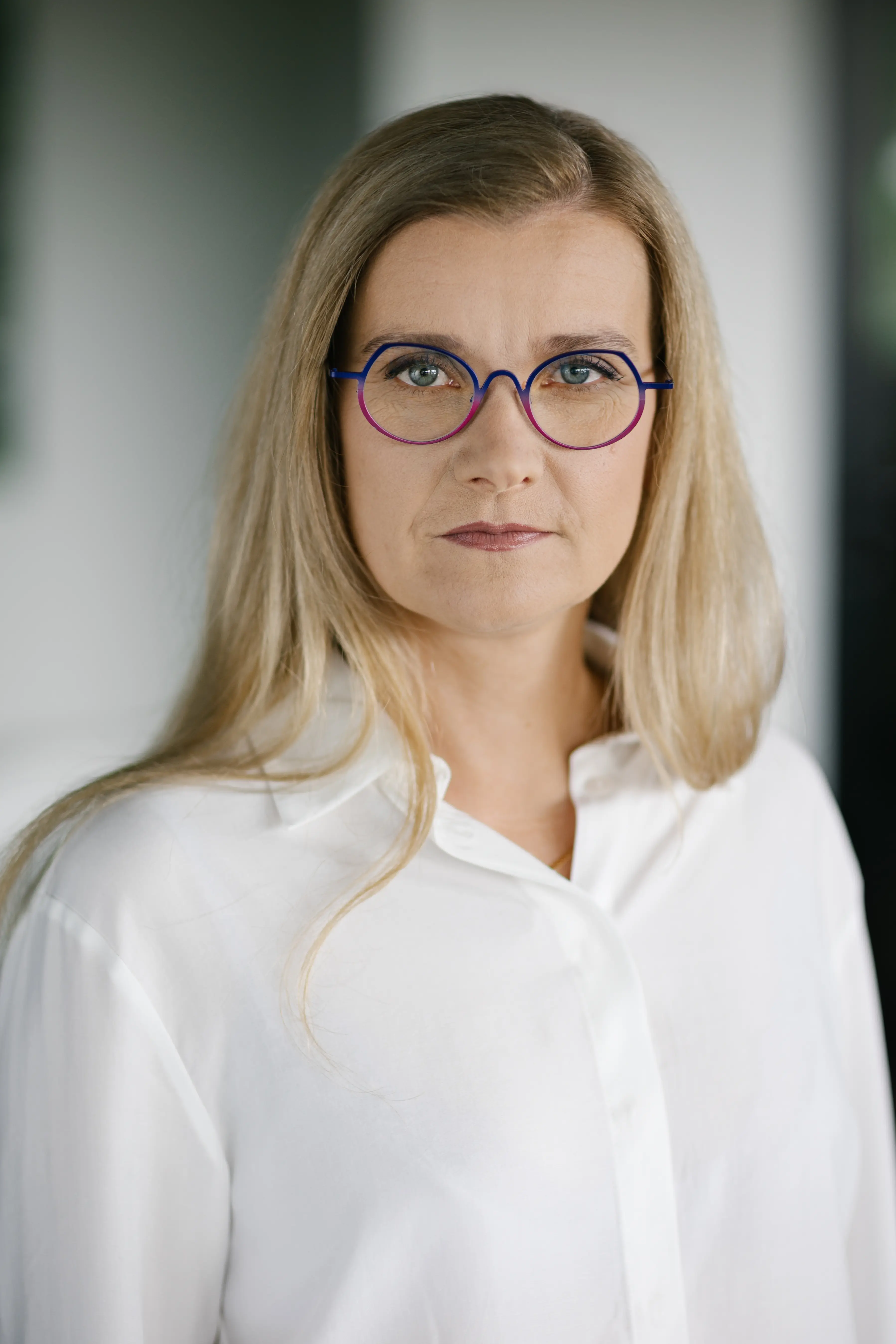 Kobieta z długimi blond włosami, ubrana w białą koszulę z kołnierzykiem i okrągłe okulary w niebieskich oprawkach, patrzy bezpośrednio w kamerę z neutralnym wyrazem twarzy. Tło jest delikatnie rozmyte.