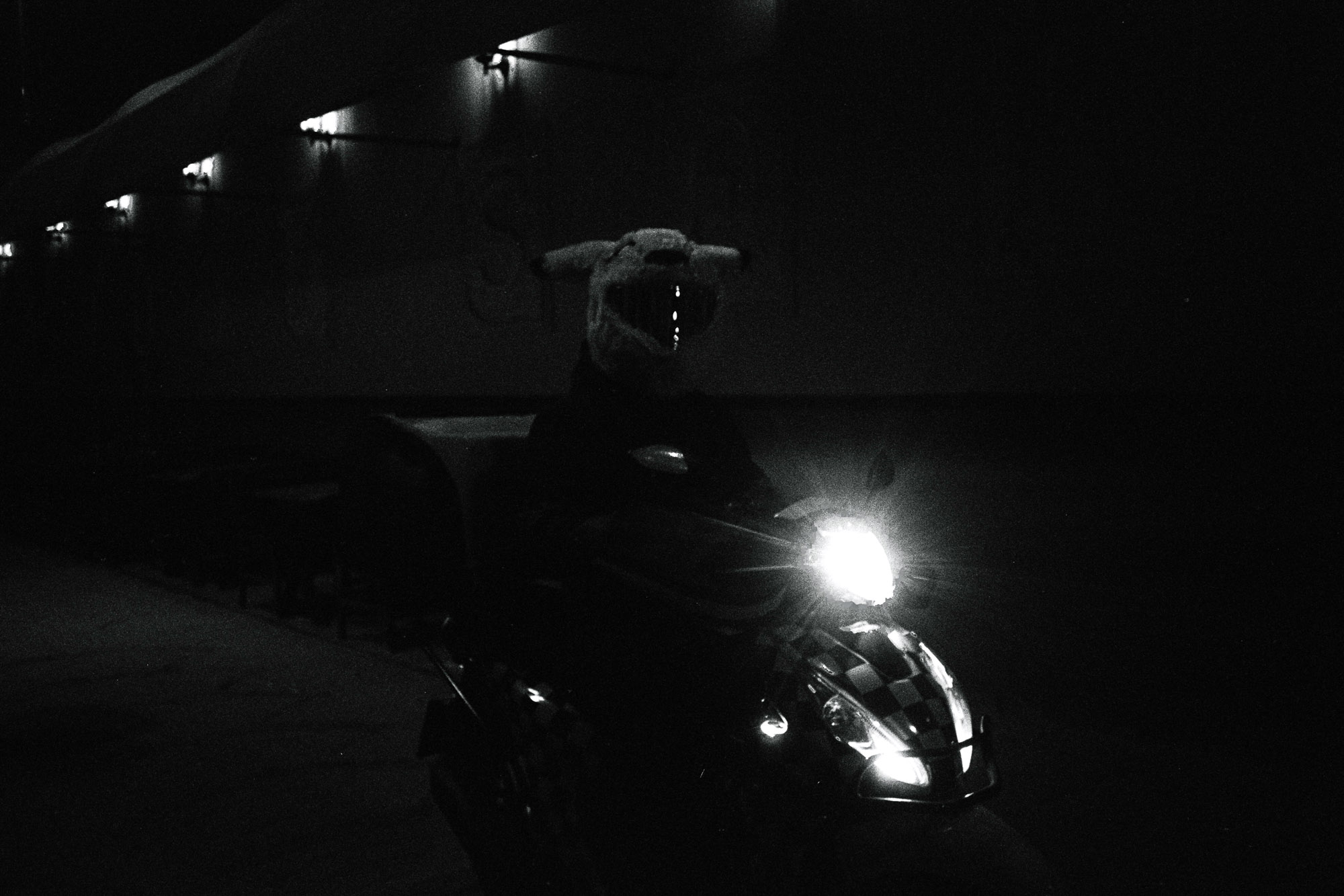 A person wearing a large, furry costume head rides a motorcycle at night, with the bike's headlight shining brightly in the dark street.