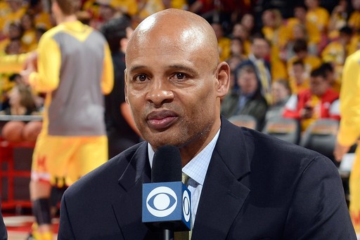 Clark Kellogg in a suit holds a CBS microphone while speaking at a basketball game. Players in yellow uniforms and spectators are visible in the blurred background.