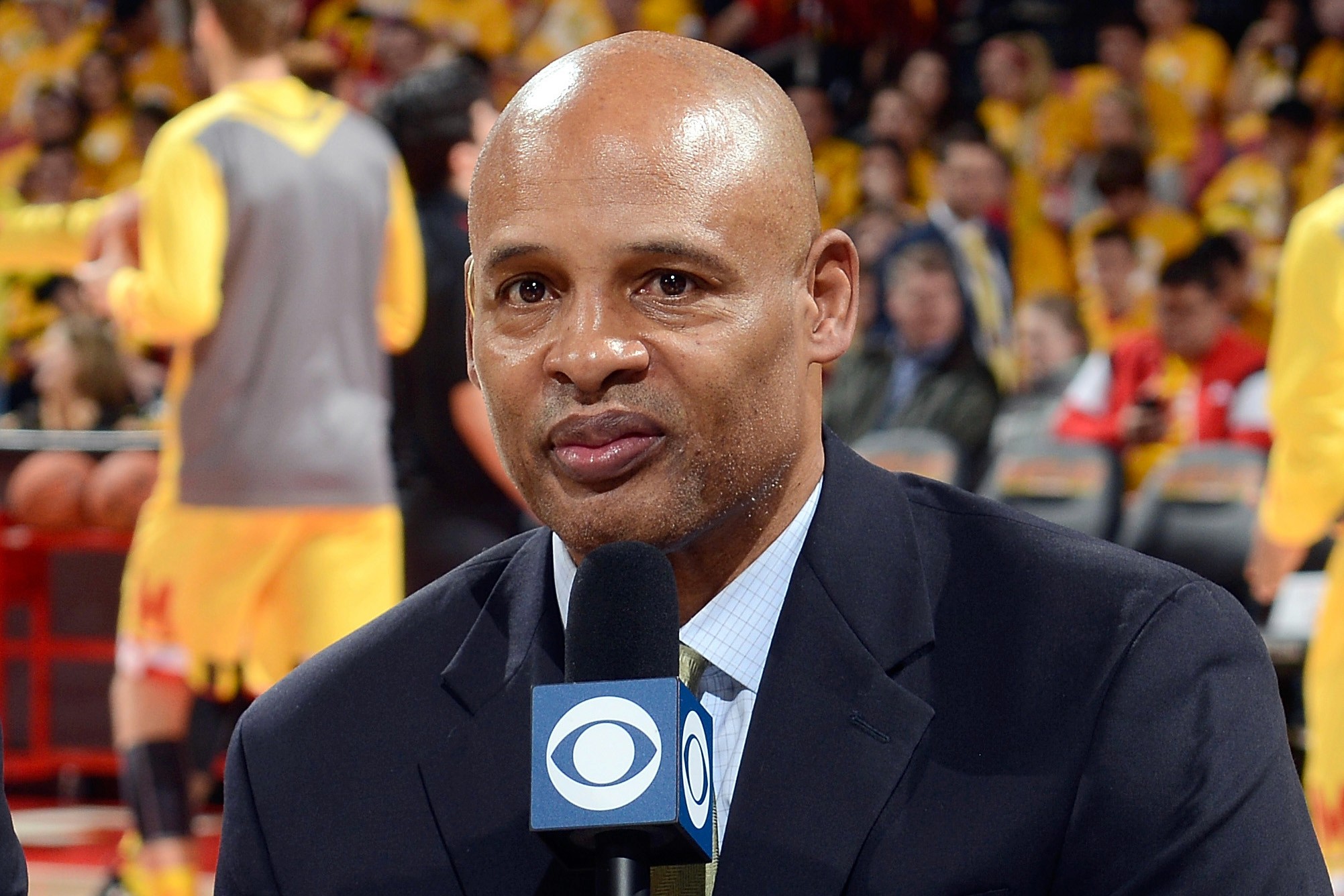 Clark Kellogg in a suit holds a CBS microphone while speaking at a basketball game. Players in yellow uniforms and spectators are visible in the blurred background.