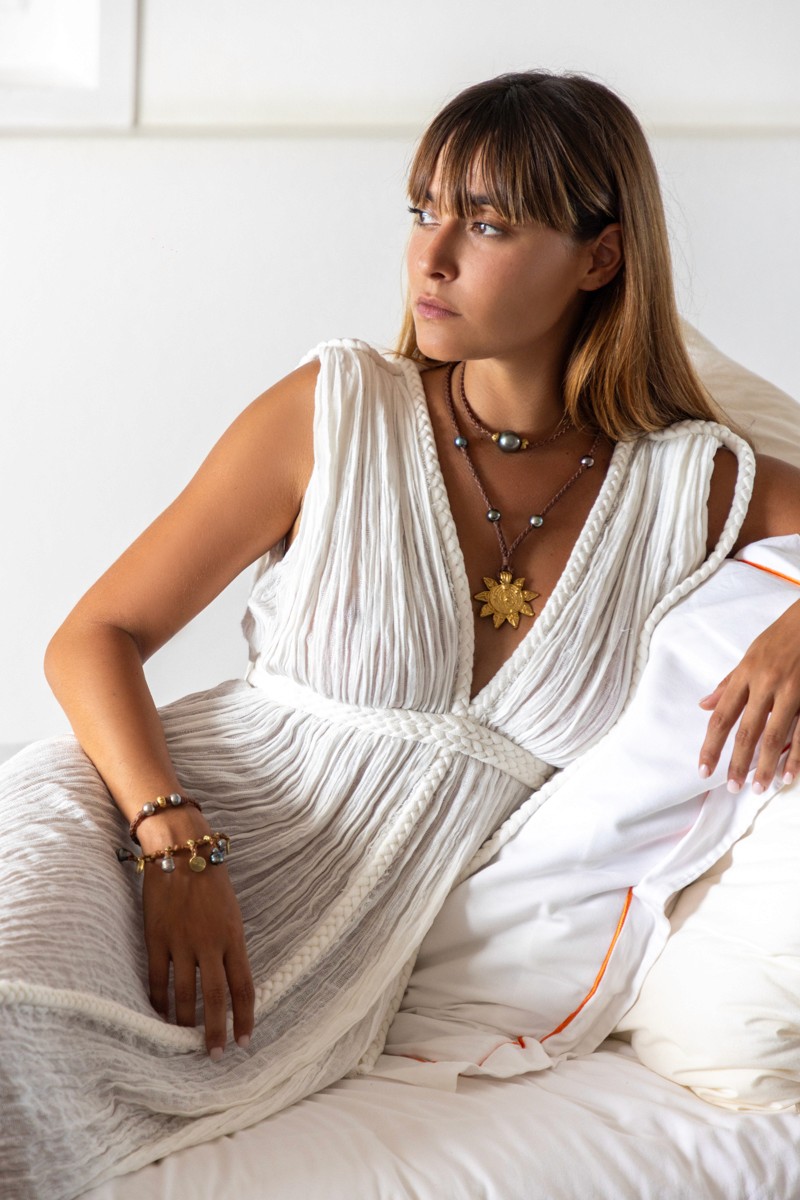 Elegant woman lounging in a white textured dress, adorned with Manos de Oro Saint Barth handcrafted luxury jewelry—featuring a radiant gold sun pendant, pearl-accented necklaces, and artisanal bracelets that embody soulful craftsmanship and island-inspired sophistication.