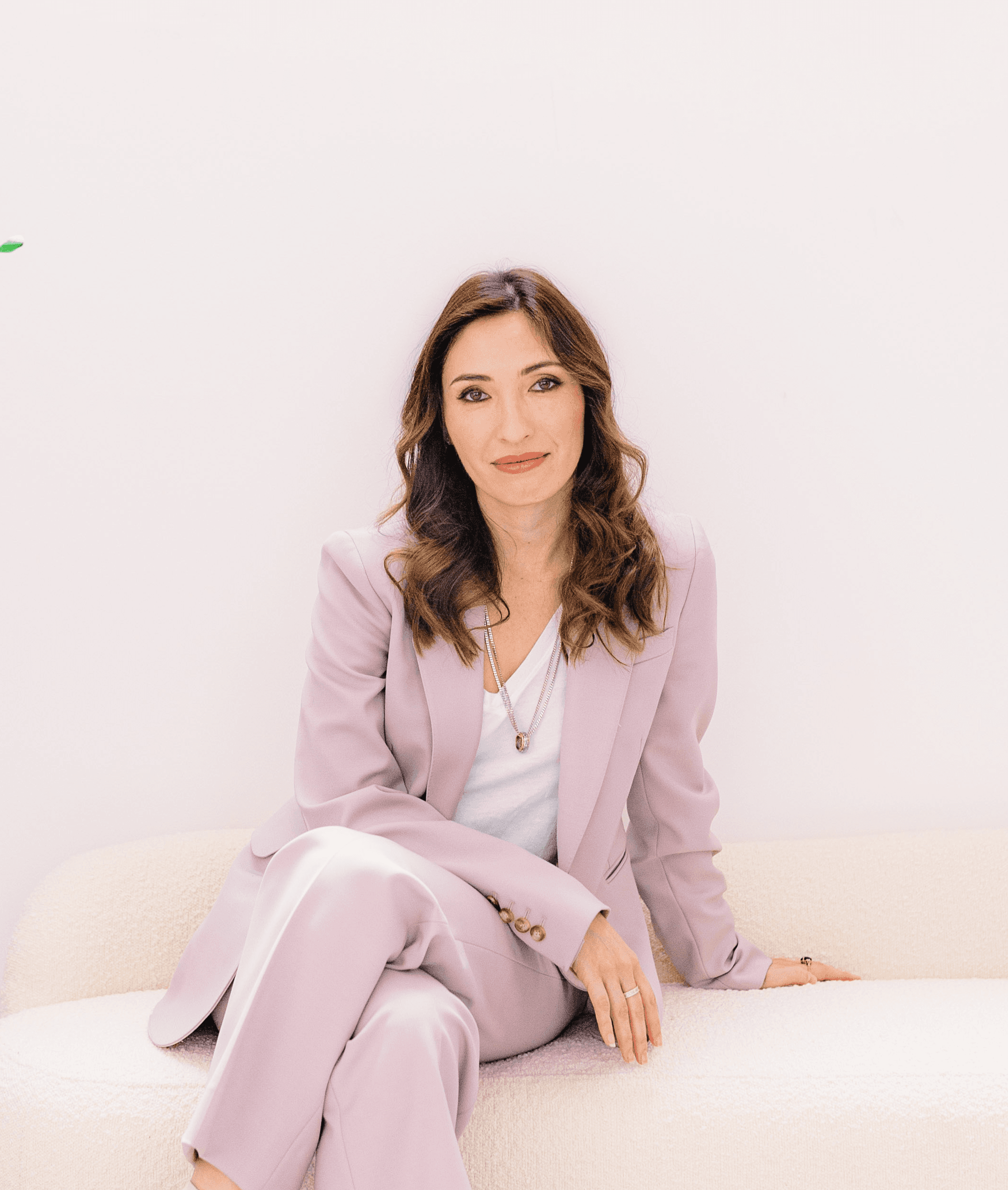 A woman in a light lavender suit sits gracefully on a cream sofa, exuding confidence and calm. A green leafy plant decorates the white, minimalist background.