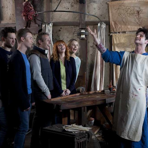 A group of five people laughing and watching a man in an apron performing in a rustic, art-studio-like setting.