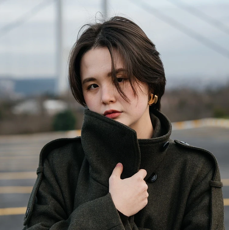 A person in a dark coat stands outdoors, holding the collar close. They have a thoughtful expression, set against a blurred, cool-toned background.